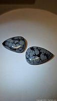 Two pear-shaped cabochon cut snowflake obsidian stones with distinctive white-gray snowflake patterns on black base, on white background