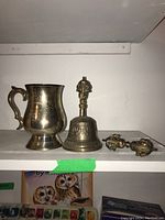 Silver-plated mug, metal bell with sculpted handle, and small decorative metal item shown together on shelf.