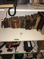 Shelf showing about 20 vintage books lined up including visible wear on spines and covers, set on a white shelf with electronic devices beneath.