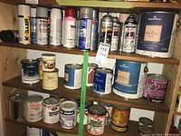 Photo of multiple shelves showing paint and chemical cans including automotive rust paint sprays, primers, and interior paint cans.