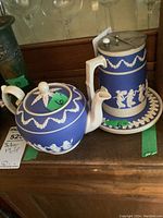 Photo showing the hot water pitcher with metal lid and matching saucer alongside the small teapot, highlighting the decorative jasperware detailing and overall condition.