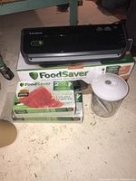 Full view of FoodSaver vacuum sealer with two rolls of heat-seal vacuum bags and vacuum sealing jar placed next to box