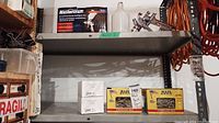 Shelf with Mastercraft spray tool on the right with attached paint cup and a glass jar next to it, above two boxes of Maze Stormguard nails and two boxes of Clous 2 1/2 nails labeled Falaise Cliffside on a metal shelving unit.