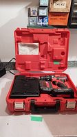 Opened hard red plastic carrying case showing Milwaukee cordless drill, battery, and manual inside case compartments.