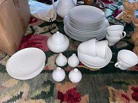 Full view of stacked dinner plates, luncheon plates, side plates, teacups stacked inside saucers, salt and pepper shakers, sugar bowl, all white porcelain with embossed patterns.