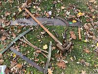 Overview of all outdoor garden tools including large bow saw, tiller, saw blades, and garden stake on leaf-covered ground.