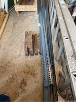 Photo showing metal corner beams with numerous perforations and three rusted metal blocks on concrete floor next to aluminum ladder.