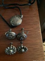 Sterling silver locket necklace and two pairs of sterling silver earrings on wooden surface. Locket is oval with engraved floral design hanging from black ribbon. One pair of earrings is floral design screw-back style, second pair engraved with raised floral pattern screw-back style.