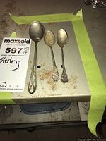 View of three sterling silver spoons arranged on a metallic surface with green masking tape