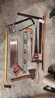 Photo showing hacksaw, bow saw, hand saw, axe, crowbar, carpenter square, spirit level, and wooden measuring sticks arranged on concrete floor.