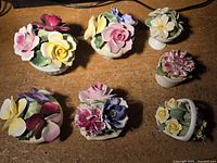 Seven small bone china floral figurines displayed on wooden surface. Various flower types including roses, pansies, carnations, and daisies. Some minor visible chips.