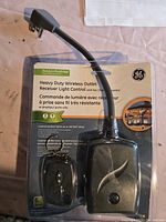 Photo of GE heavy duty wireless outlet receiver light control in packaging showing front of receiver and attached power plug. Black plastic device with round button and antenna wire.