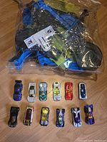 Plastic race track components in a sealed clear bag with 12 Hot Wheels cars arranged in front on wooden floor
