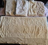 Photo showing a large cream-colored knitted or crocheted runner with intricate geometric patterns and fringed edges, accompanied by several smaller linens folded above it.