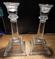 Pair of clear glass candleholders on wooden surface with black background, showing stepped square base, faceted column with rings, and flared cup top.