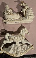 Top-down view of two white and gold ceramic Christmas figurines: Santa in sleigh with reindeer and rocking horse with gifts and wreath underneath.