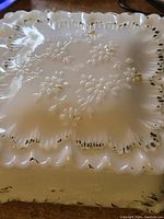 Top view of square white milk glass trinket box lid showing floral embossed pattern and scalloped edge with black paint accents worn.