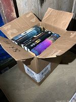 Opened cardboard box showing a variety of novels inside, mostly paperbacks and one hardcover, arranged standing vertically.