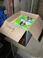 Box labeled 'Cooking' filled with cookbooks shown partially inside a cardboard box in a dimly lit area