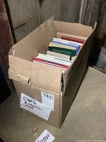 Photo of a cardboard box containing approximately 18 books of assorted sizes and genres.