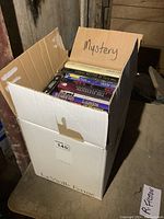 Photo showing the box labeled 'Mystery' full of novels stacked vertically inside a cardboard box on a wooden floor.