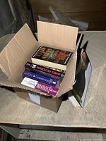 Box partially open on a mattress containing five visible novels with varied cover designs.