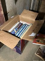 Open cardboard box filled with assorted books seen from top-down angle.