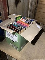 Box of books with black, colorful, and white spines, including mystery titles and novels by Tom Clancy, Dean R. Koontz, and others.