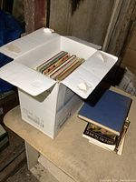 Box filled with assorted books on a small table, books stacked beside the box