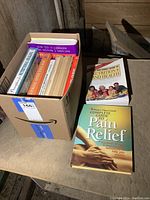 Box showing several self-help books, including 'The Complete Guide to Pain Relief' and 'The Family Guide to Nutrition and Health' on top of the box, with more books inside.