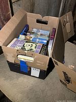 Box of assorted novels and books showing partial titles and spines