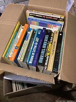 Box full of various books, mixed genres, showing titles like The Stressed Balance, Betty Friedan's The Second Stage, Art Now, and others