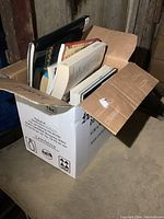 Box full of mixed genre books including cookbooks and fiction stacked inside a cardboard box.