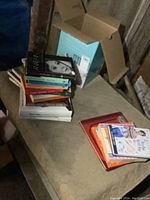 Box with stacked novels and kids books, some titles visible on top such as The Diary of Anais Nin and Steve Jobs.