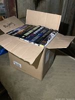 Box full of hardcover novels, viewed at an angle showing many books packed vertically inside a cardboard box with visible wear on the box.