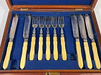Full set of flatware in blue velvet lined wooden box showing five forks and five knives with cream colored handles.