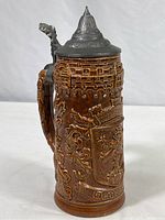 Full view of ceramic beer stein with pewter hinged lid showing handle and decorative relief.