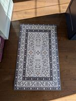 Photo showing the full rectangular anti-fatigue mat laid on a wooden floor with natural light. The mat features a patterned border with floral motifs in grey and beige on a light base color.