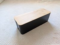 Black rectangular box with wooden lid placed on carpet floor, showing overall external appearance and size.
