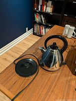 Cordless kettle and its separate power base seen on wooden table with bookshelves in the background.