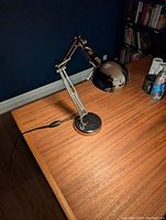 Chrome adjustable desk lamp on wooden desk, showing the lamp on with power cord positioned on table with inline switch visible.