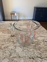 Front and side view of clear glass Fire King measuring jug with red measurement markings and sturdy handle, 1 liter capacity.