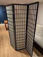 Three-panel black wood framed folding screen with white paper panels standing on wood floor, angled view showing entire screen.