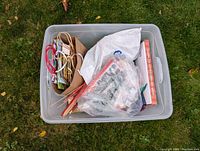 Plastic storage bin filled with paper gift bags, strung decorations, and a bag containing wooden sticks and other holiday decoration items.
