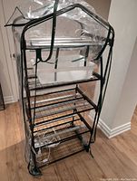 Full view of the indoor plant nursery rack showing four wire mesh shelves, clear plastic greenhouse cover with zippered front, and a white tray on second shelf.
