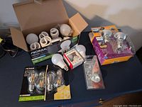 Lot of various light bulbs displayed on a dark surface with some in original boxes and others loose, including spiral CFLs, round LED bulbs, and reflector bulbs.