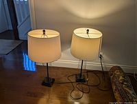 Two matching lamps standing on a wooden floor near a wall. Lamps have beige fabric shades and cylindrical glass bases mounted on square black supports. Power cords visible loose on the floor.