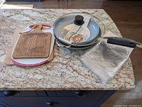 Photo showing two cutting boards, cork trivet and transparent plastic table cloth next to the three pans with glass lid on a granite countertop.