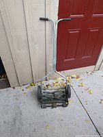 Photo of a manual push reel lawn mower standing against a beige shed wall with a red door to the right. The mower has a metal frame, two large wheels, and a U-shaped handle with black grips.