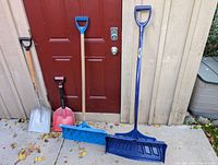 Four shovels standing against a wooden wall with a red door in the background. Two snow shovels with blue blades and two regular shovels with metal blades.